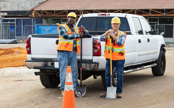 two construction workers smiling behind their white work truck with shovels
